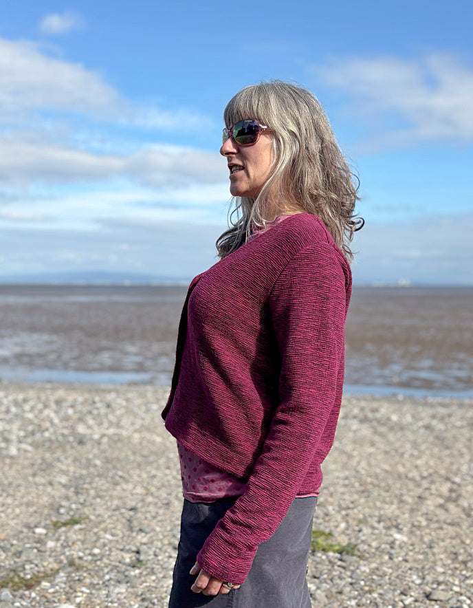 Woman wearing a maroon sweater standing on a beach with a blue sky and clouds in the background.
