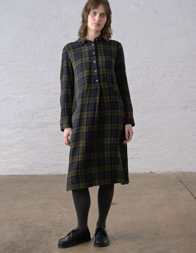 A woman stands against a white textured wall wearing a long-sleeve plaid shirt dress in dark green, black, and brown tones, paired with black tights and black shoes.