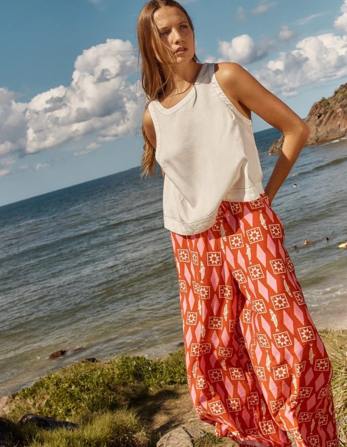 Woman in a white sleeveless top and red patterned pants standing on a beach with ocean and sky in the background.