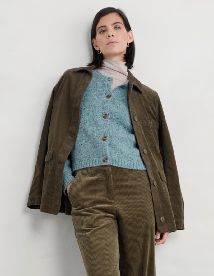 Person wearing a brown jacket, blue knitted cardigan, and brown pants against a white background