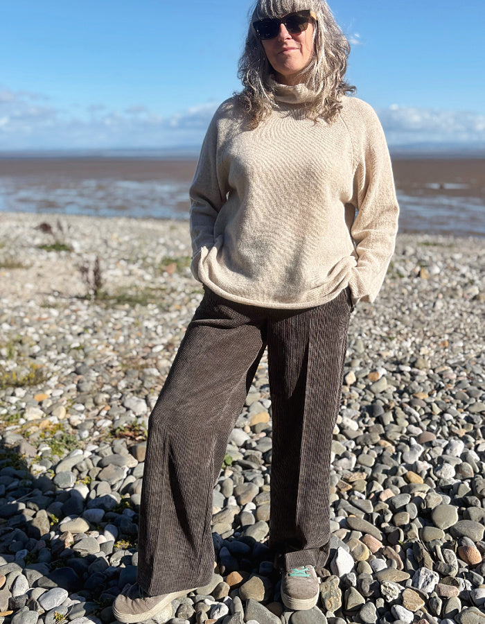A woman standing on a pebble beach wearing dark brown wide-leg corduroy pants, a cream knit sweater, and dark sneakers, with water and sky in the background.