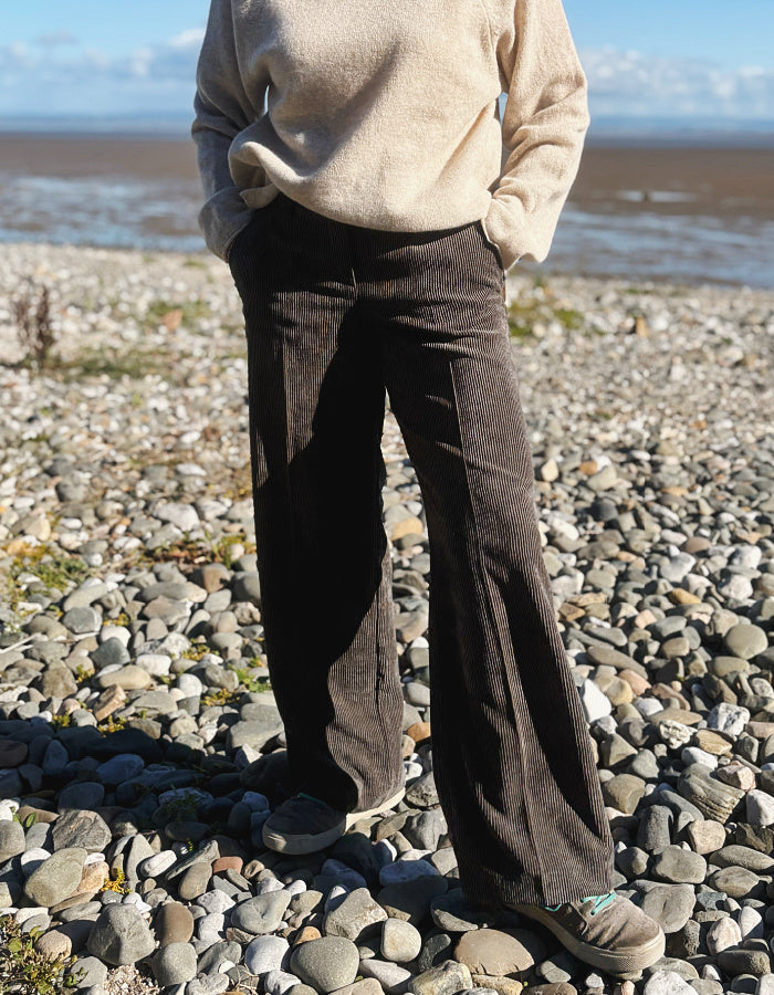 A woman standing on a pebble beach wearing dark brown wide-leg corduroy pants, a cream knit sweater, and dark sneakers, with water and sky in the background.
