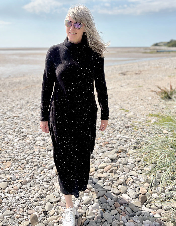 Person wearing a black dress with white speckled galactic print standing on a pebbly beach with a blue sky.