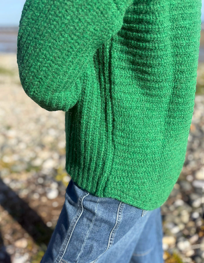 A close-up of a person wearing a bright green ribbed knit sweater and blue jeans outdoors on a pebble beach.