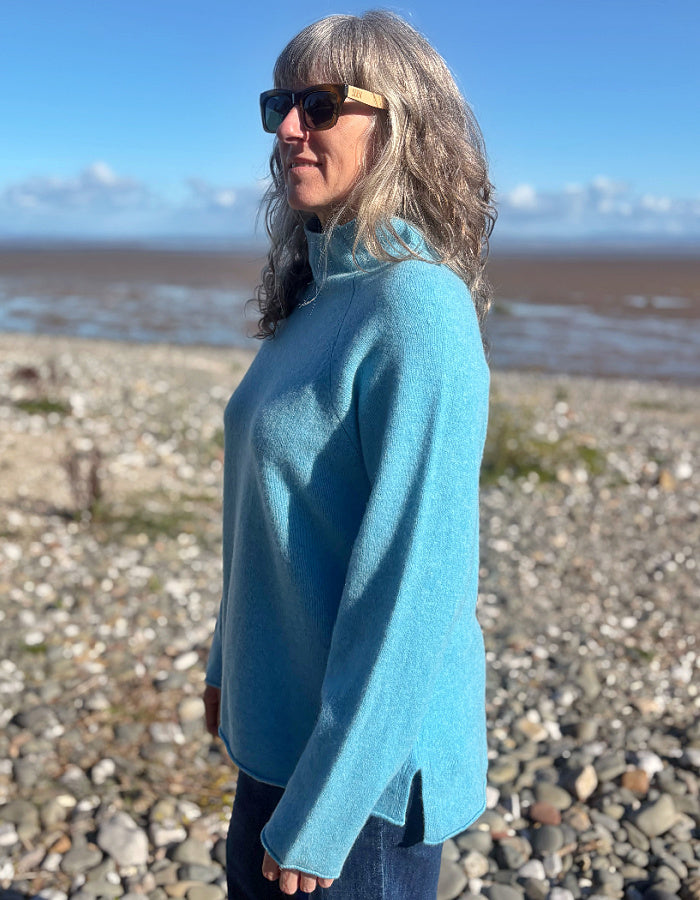 A woman with gray hair and sunglasses wearing a bright turquoise blue mock-neck sweater, standing on a pebble beach with the sea and blue sky in the background.