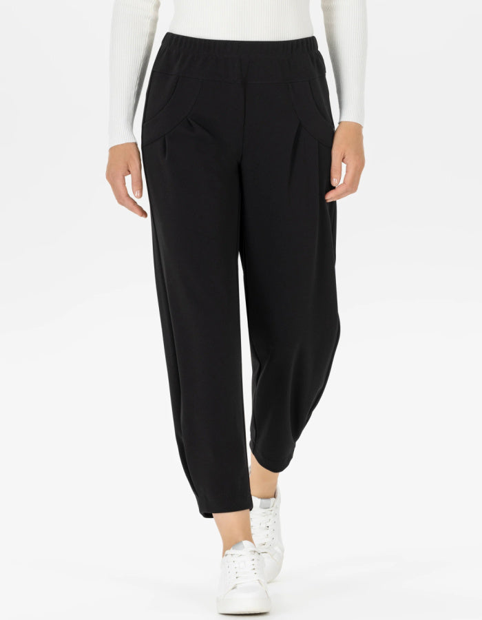 A person wearing black cropped pants with an elastic waistband, side pockets, and a loose tapered fit, styled with a white long-sleeve top and white sneakers against a light gray studio background.