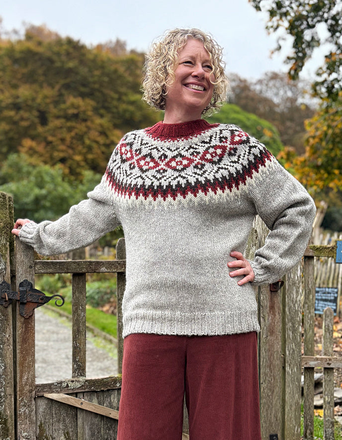 Woman wearing a patterned sweater standing in front of a wooden gate with trees in the background