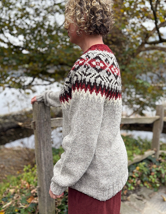 Person wearing a patterned sweater standing outdoors with trees and water in the background