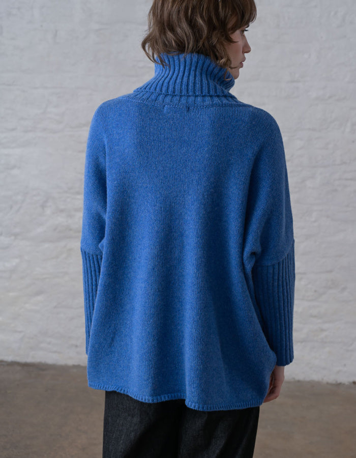A person shown from the back wearing a bright blue ribbed turtleneck sweater with ribbed elbow patches, paired with dark pants, standing against a white textured wall.