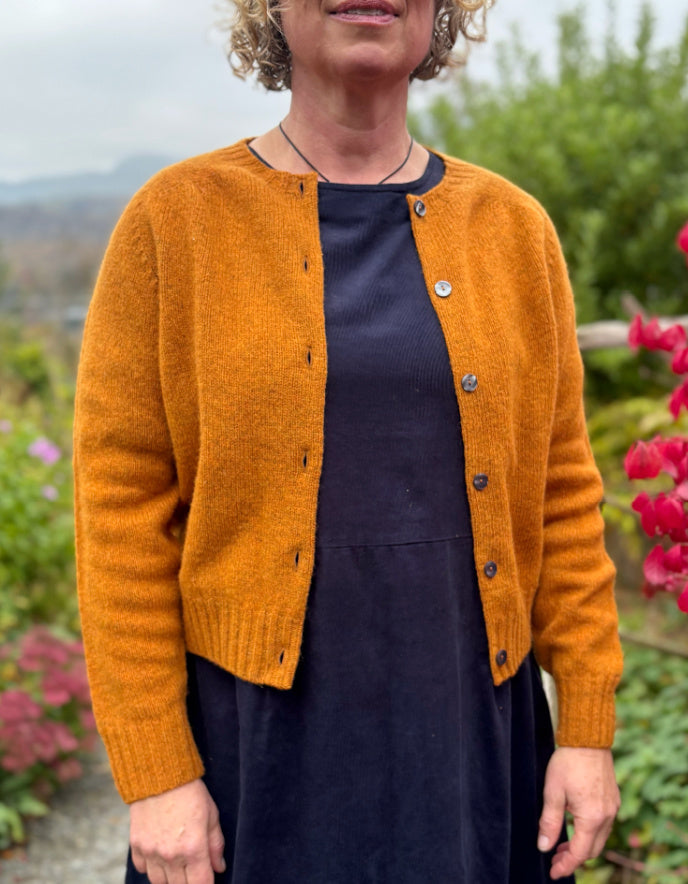 Person wearing a mustard yellow cardigan over a dark dress, standing outdoors with greenery and flowers in the background.