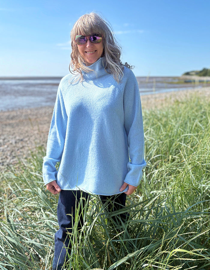Person wearing a light blue sweater standing in tall grass by a beach.