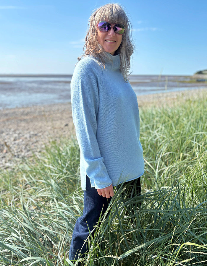 Person wearing a light blue sweater standing in tall grass by a coastal area