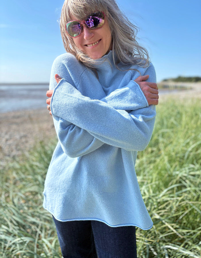 Person wearing a light blue sweater standing on a beach with grass and water in the background