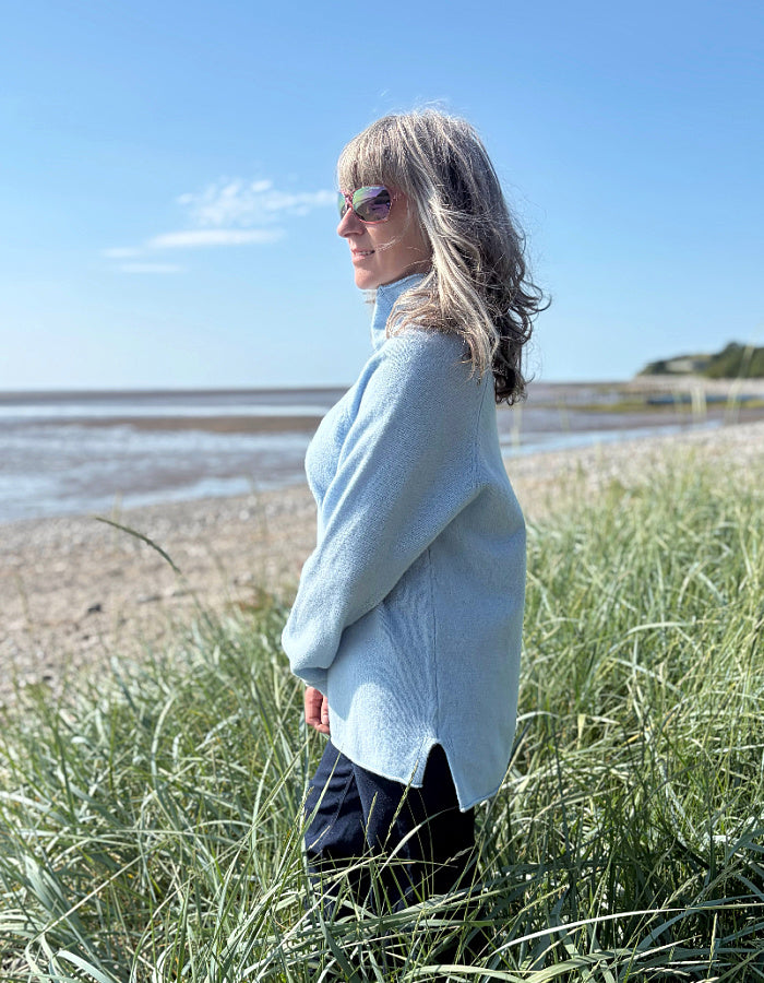 Woman in a light blue sweater standing on a beach with grass in the foreground