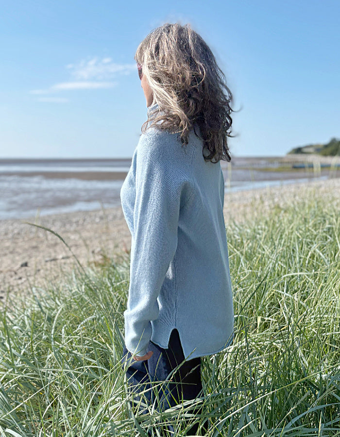 Person standing on a beach wearing a light blue wool sweater, looking out towards the water.