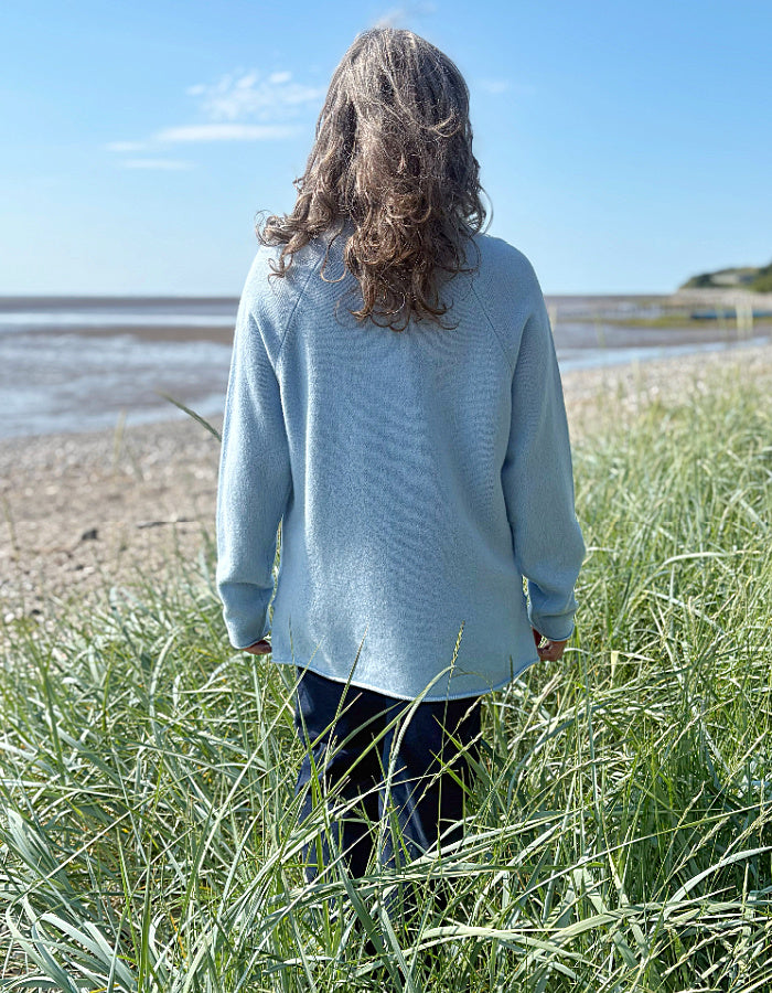 Person standing on a beach wearing a light blue wool sweater, looking out towards the water.