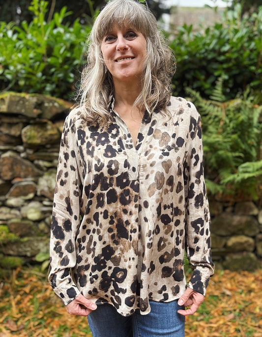 Woman wearing a leopard print shirt outdoors with greenery and a stone wall in the background