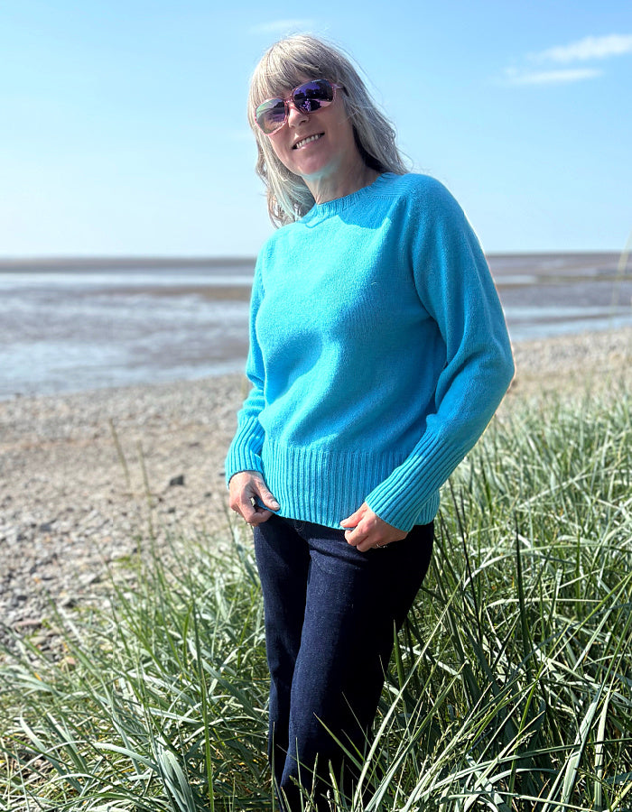 Person wearing a bright blue sweater standing on a beach with grass in the foreground.