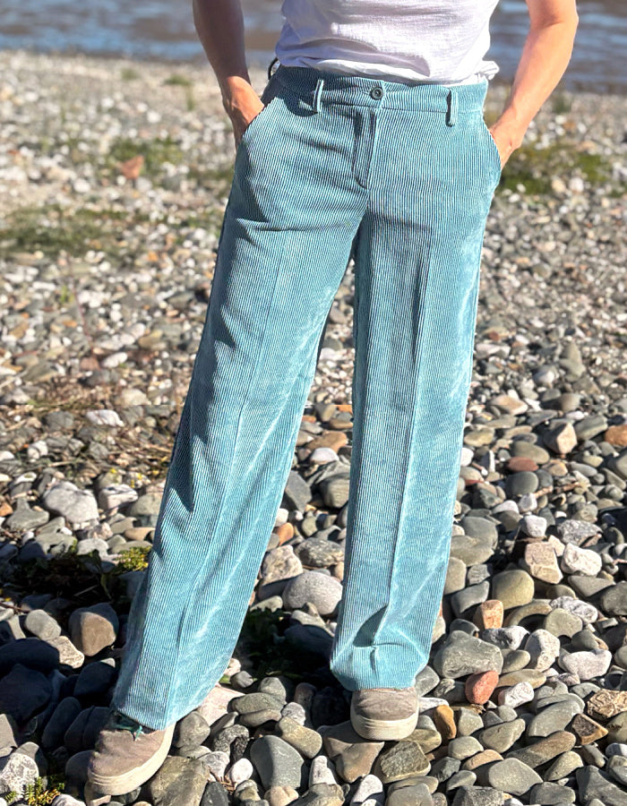 A person standing on a pebble beach wearing light blue wide-leg corduroy trousers, a white t-shirt, and beige sneakers.