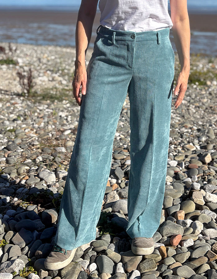 A person standing on a pebble beach wearing wide-leg teal corduroy trousers, a white top, and beige sneakers.