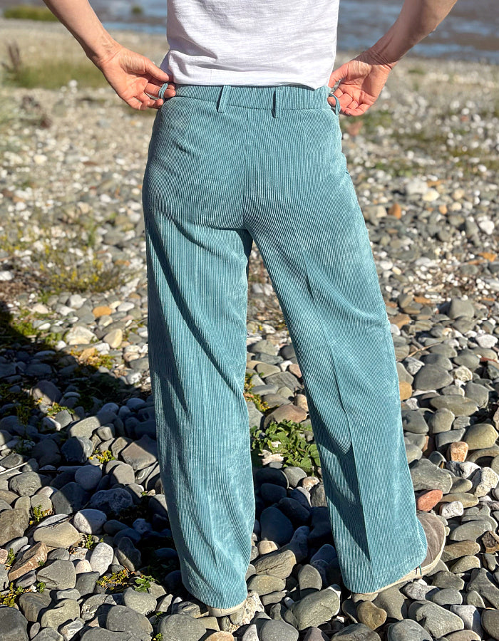 A person standing outdoors on a pebble beach wearing wide-leg corduroy trousers in a muted teal-blue color, paired with a white top. The photo is taken from behind, showing the back of the pants clearly.