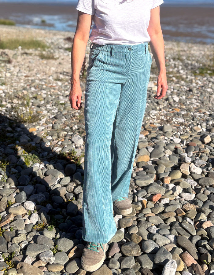A person standing on a pebble beach wearing light blue wide-leg corduroy pants, a white t-shirt, and brown shoes.