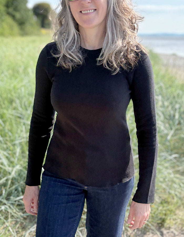 Woman wearing a black long-sleeve top and blue jeans standing in a grassy field with a blurred background