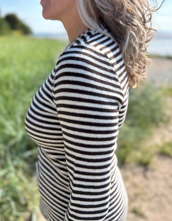Woman wearing a black and white striped long-sleeve shirt standing outdoors with greenery and a body of water in the background.