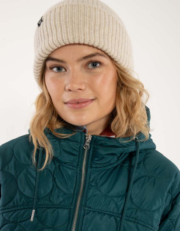 Person wearing a beige knit beanie and teal quilted jacket on a white background