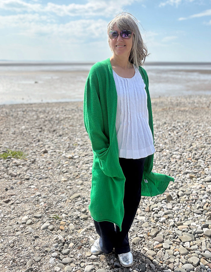 Person wearing a green wrap long merino wool cardigan on a pebbly beach with ocean and sky in the background