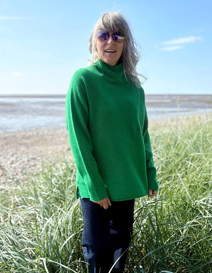 Woman wearing a green wool fisherman style sweater and sunglasses standing on a grassy field with a beach in the background.