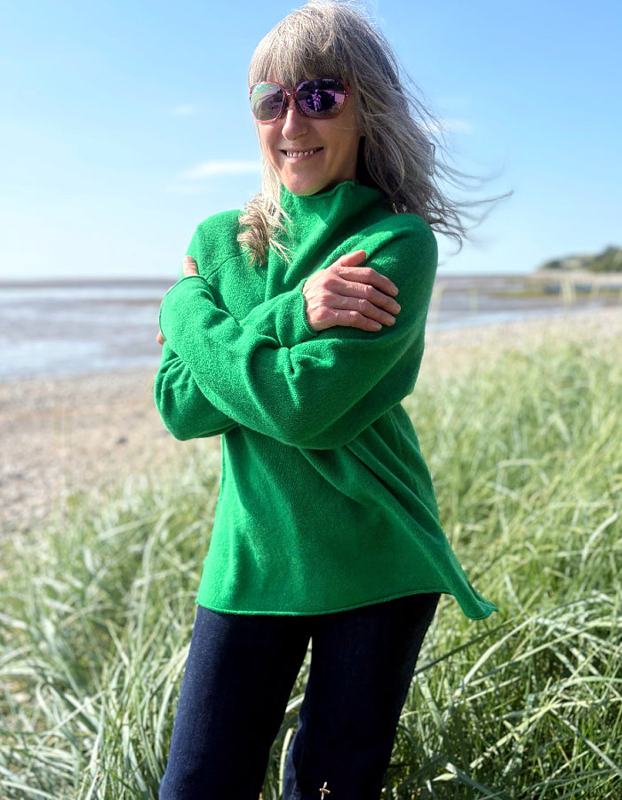 Woman wearing a green wool fisherman style sweater and sunglasses standing on a grassy field with a beach in the background.