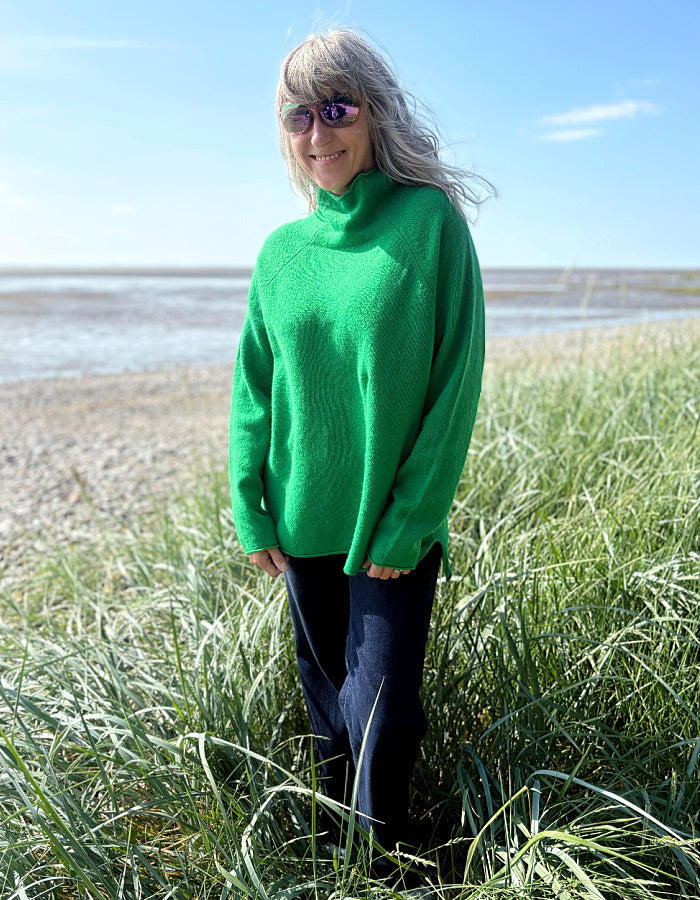 Woman wearing a green wool fisherman style sweater and sunglasses standing on a grassy field with a beach in the background.