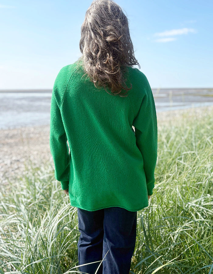 Woman wearing a green wool fisherman style sweater and sunglasses standing on a grassy field with a beach in the background.