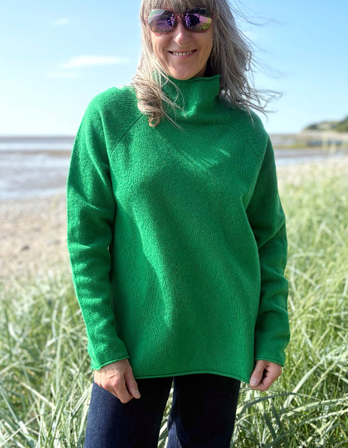 Woman wearing a green wool fisherman style sweater and sunglasses standing on a grassy field with a beach in the background.