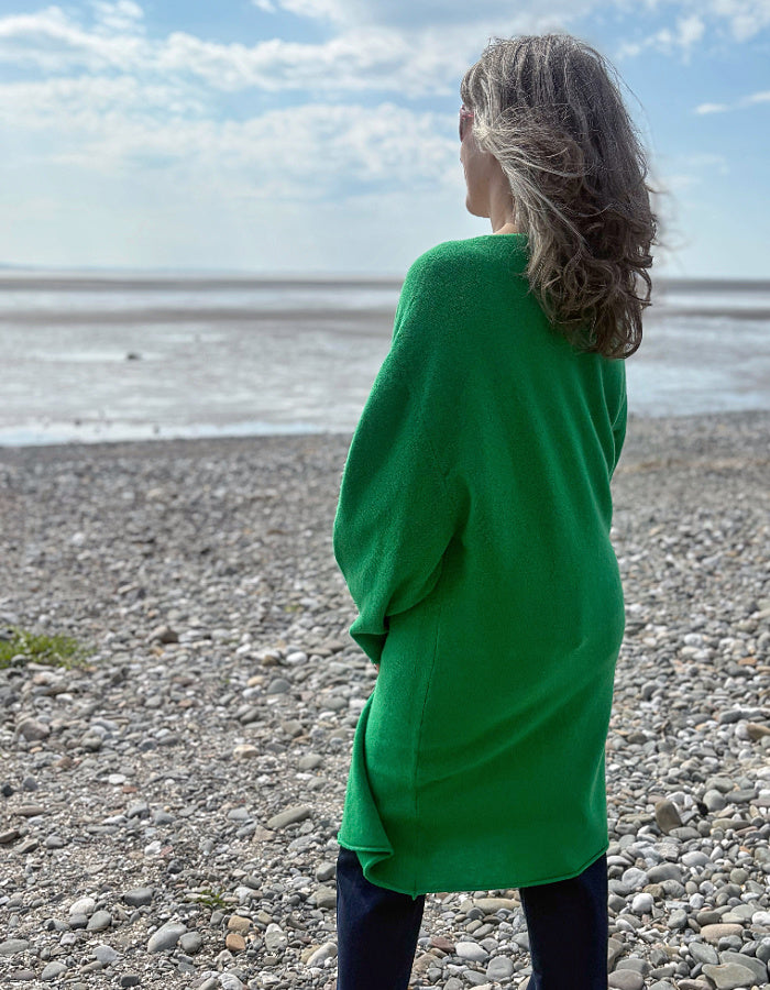 Person wearing a green wrap long merino wool cardigan on a pebbly beach with ocean and sky in the background