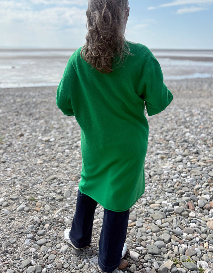 Person wearing a green wrap long merino wool cardigan on a pebbly beach with ocean and sky in the background