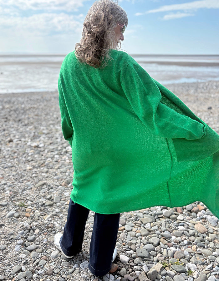 Person wearing a green wrap long merino wool cardigan on a pebbly beach with ocean and sky in the background