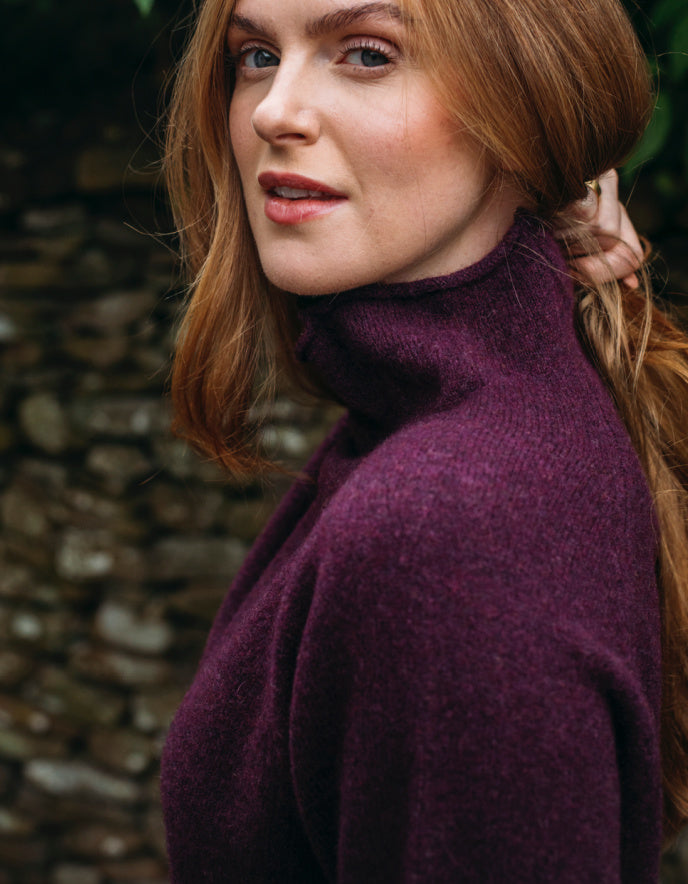 Woman wearing burgundy wool turtle neck sweater standing in front of a stone wall and greenery