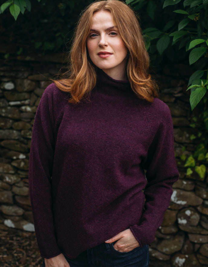 Woman wearing burgundy wool turtle neck sweater standing in front of a stone wall and greenery