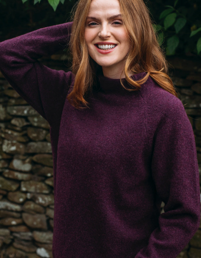 Woman wearing burgundy wool turtle neck sweater standing in front of a stone wall and greenery