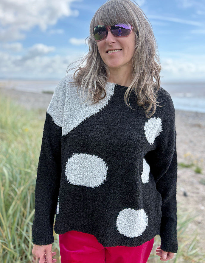 Woman wearing a black sweater with white polka dots and pink velvet pants standing on a beach.