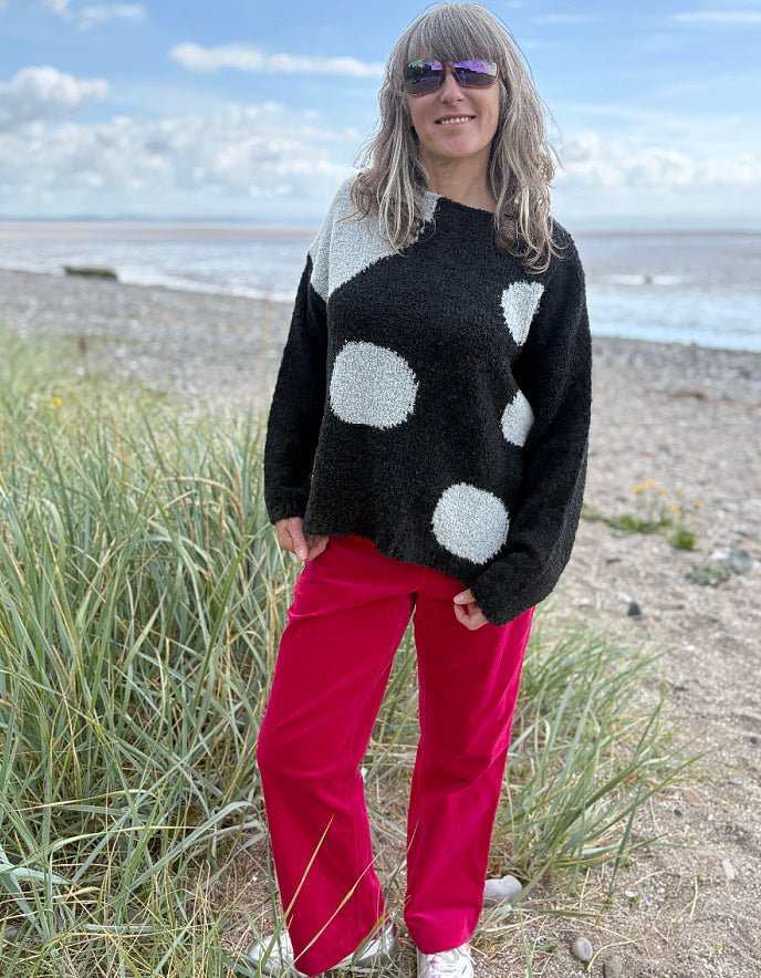 Woman wearing a black sweater with white polka dots and pink velvet pants standing on a beach.