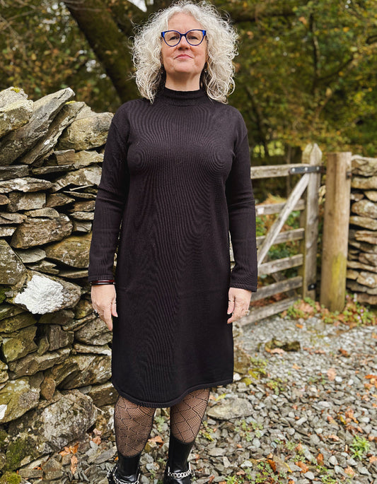 Woman wearing a black knit dress standing in front of a stone wall and wooden gate.