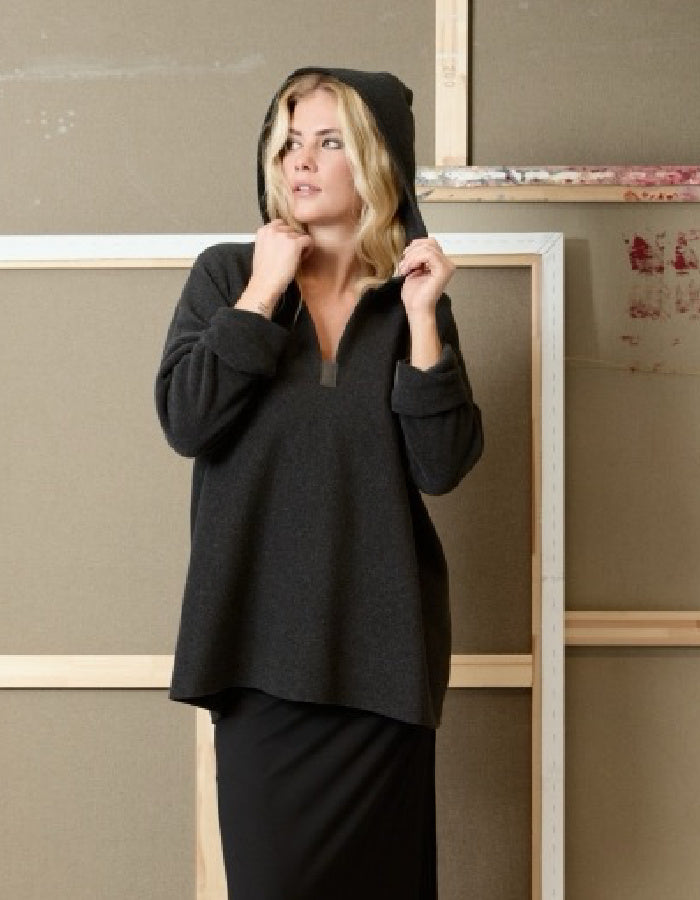 A woman wearing a dark charcoal gray pullover hoodie with a half-zip neckline and a matching black skirt, standing in front of a rustic wooden frame.