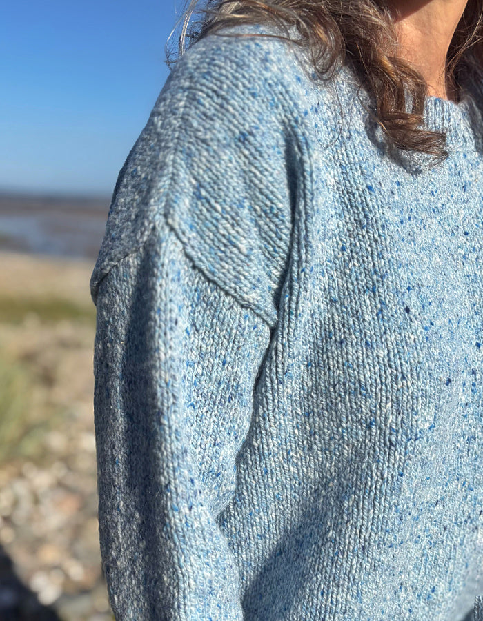 Close-up of a person wearing a light blue heathered knit sweater outdoors near a beach, with the person's shoulder, hair, and part of the neck visible.