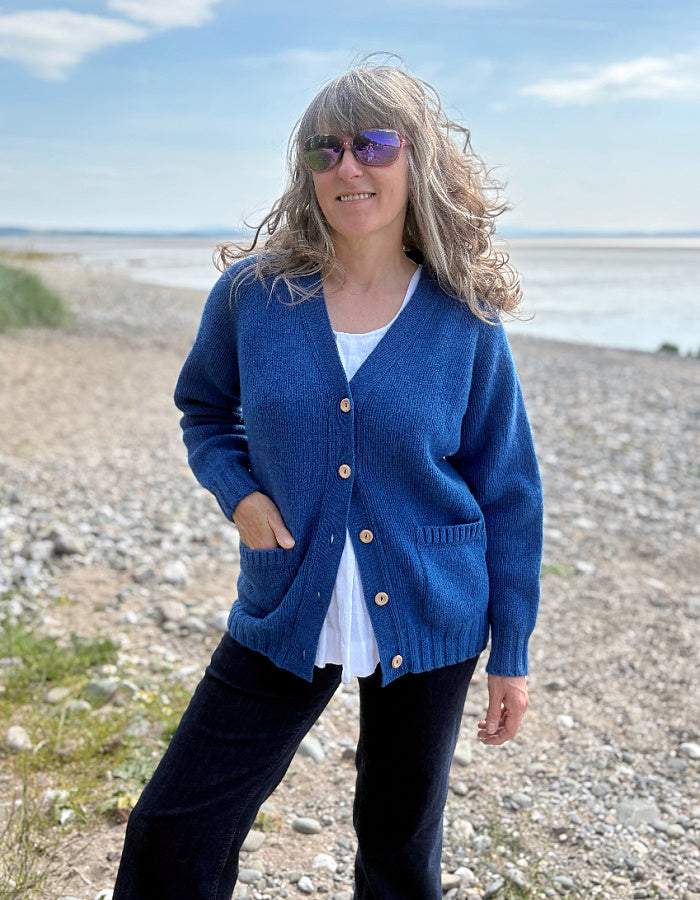 Woman wearing a wool blue cardigan on a pebbly beach with water and sky in the background