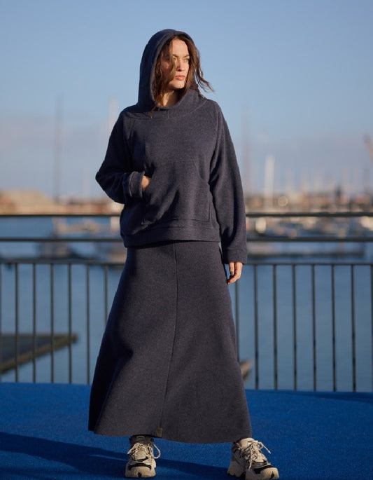 A woman wearing a dark charcoal gray two-piece set consisting of a pullover hoodie and a long A-line skirt, standing outdoors on a blue surface with a railing and water in the background.