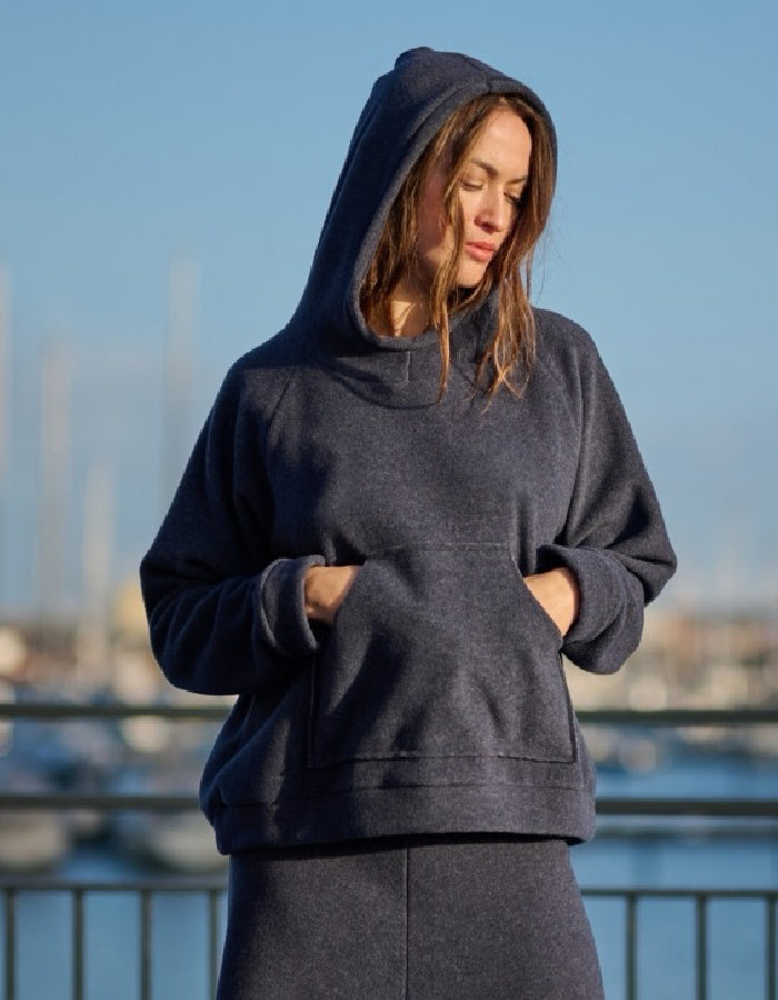 A woman wearing a dark navy blue heathered pullover hoodie with a matching sweatpants set, standing outdoors with a blurred background of water and boats.