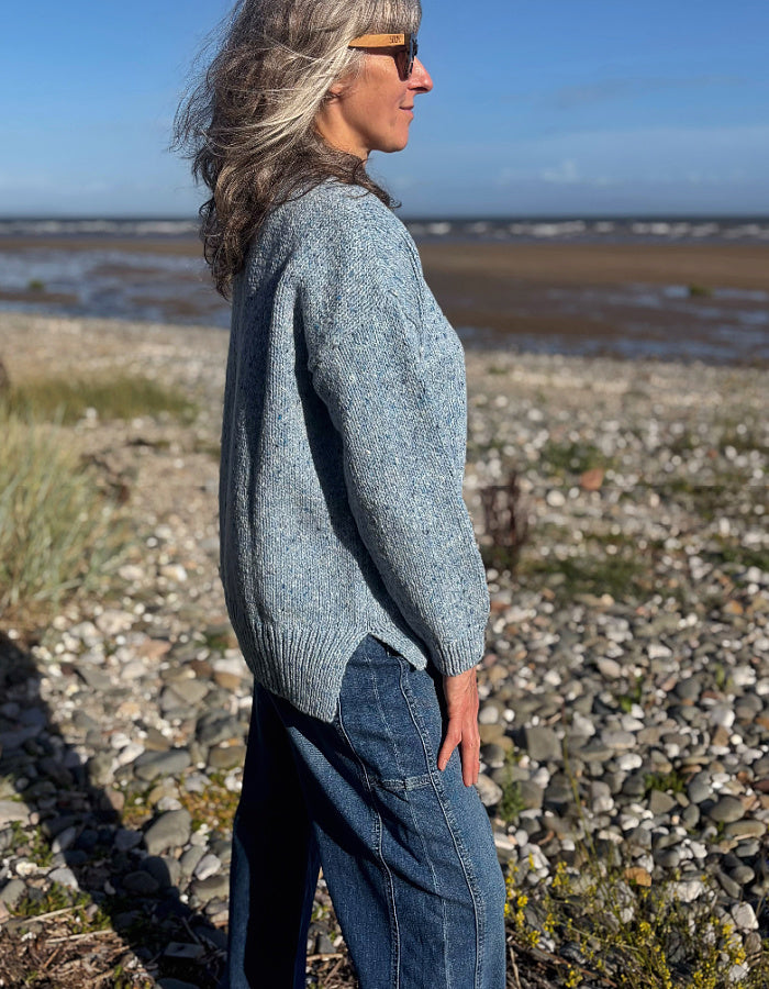 A woman wearing a light blue donegal tweed merino knit sweater and blue jeans stands on a rocky beach with water and sky in the background.